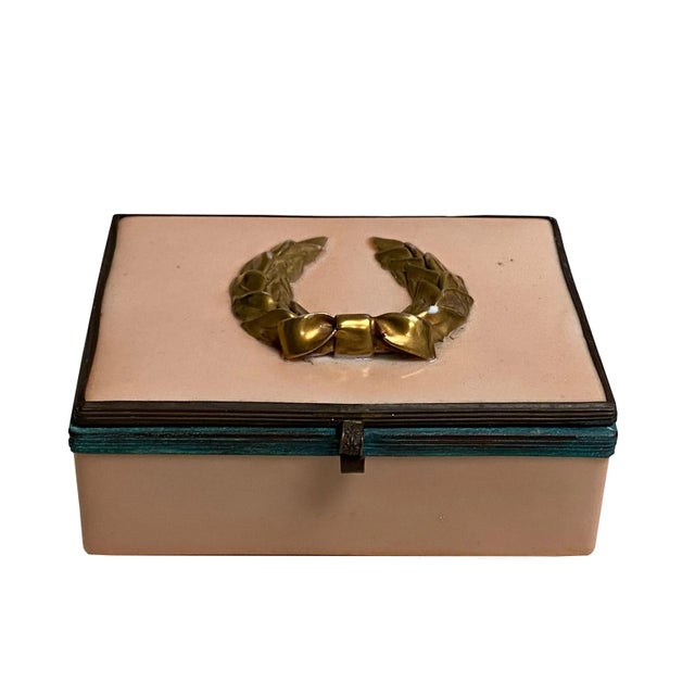 A salmon colored enameled box with patina bronze surround and latch and topped with a gold laurel wreath.