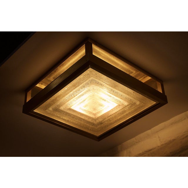 Square ceiling light in brushed and polished brass, holding a thick slab of translucent murano glass with geometric...