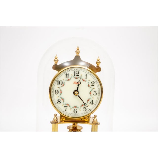 A brass German 400-day anniversary clock under a glass dome with a spired brass finial crown, white enamel dial with...