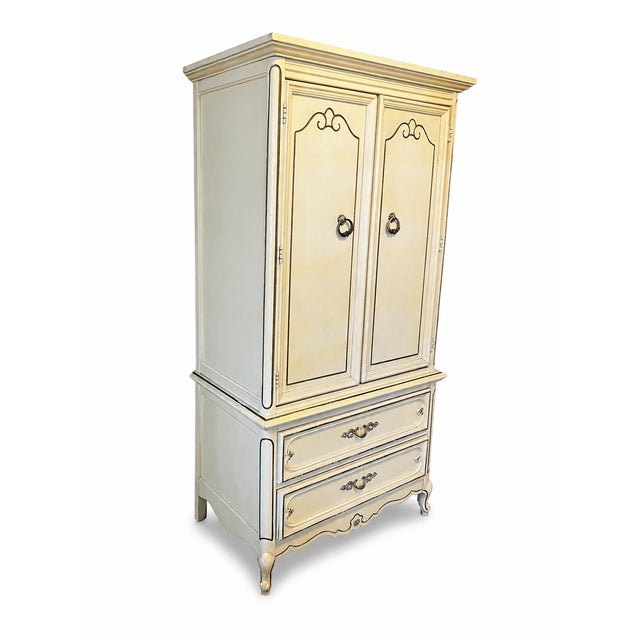 An elegant vintage French Provincial style armoire or linen press featuring a warm cream- painted finish with contrasting...