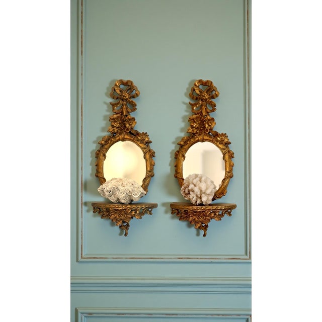 A charming pair of vintage Italian Florentine-style gilt wall shelves with integrated oval mirrors and elaborate ribbon-...