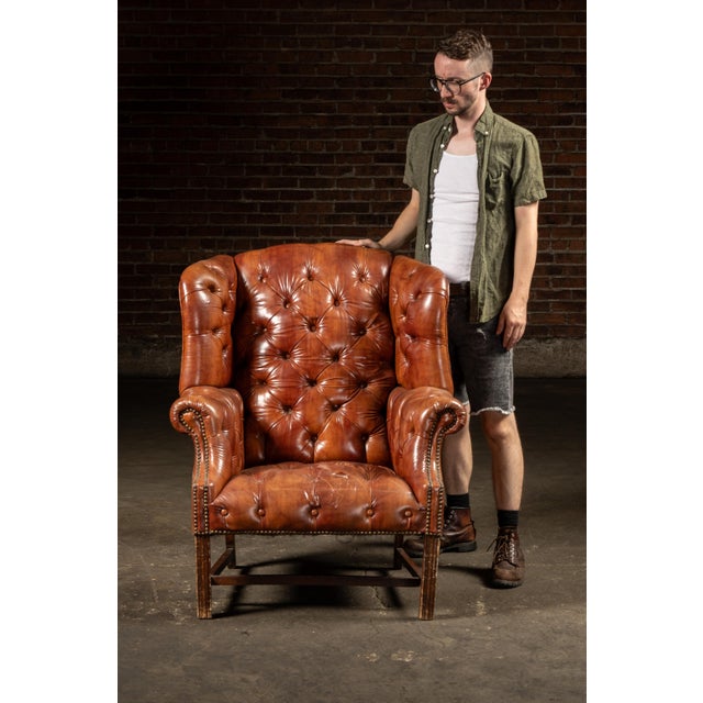 Large Chesterfield wingback chair upholstered in tufted brown vinyl over a hardwood frame. The design combines the deep...