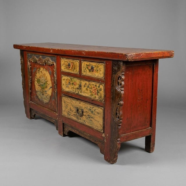 A richly weathered Tibetan cabinet console featuring hand-painted floral and figurative panels in subdued cinnabar red,...