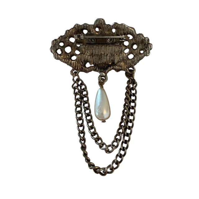 Victorian-style brooch featuring a large oval faux pearl, an ornate filigree design, and draped chains with a dangling...
