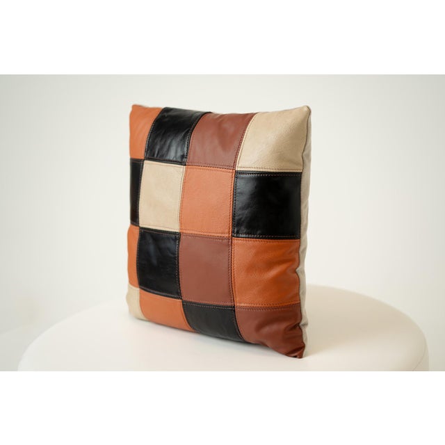 Bold and artisanal, this 16-inch square throw pillow showcases a patchwork checkerboard of orange, black, and cream...