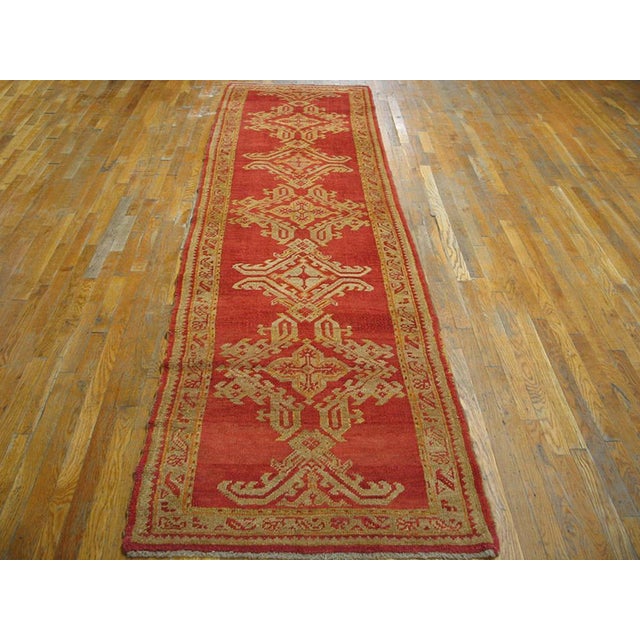 The clear red field of this circa 1900 Oushak runner displays a column of five complete and two fractional lozenge and...