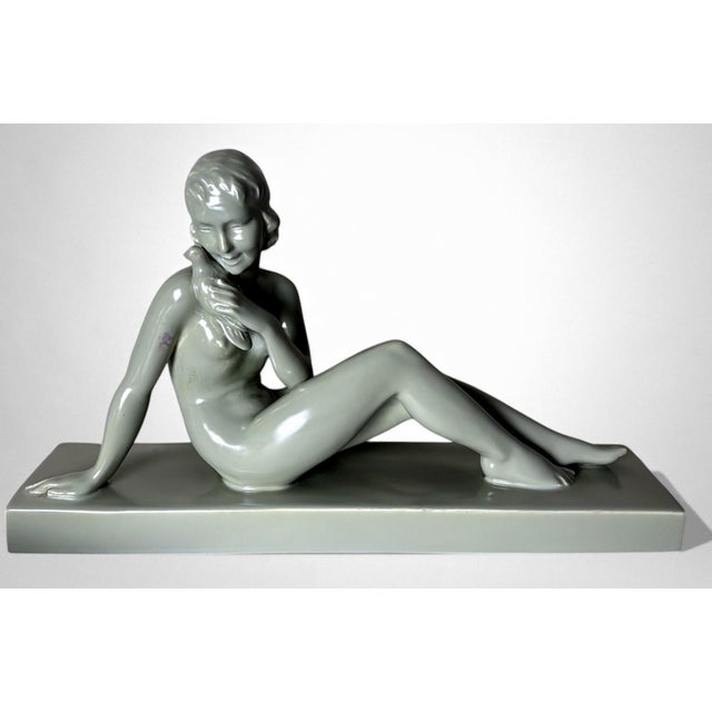 Elegant glazed ceramic sculpture depicting a nude young woman seated on an elongated base. Her composed posture, soft...