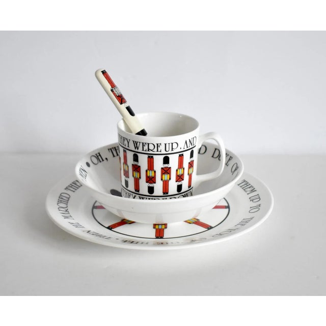 A vintage child's dining set of china, including a cup, bowl, plate, and spoon, with British soldiers in red uniforms and...