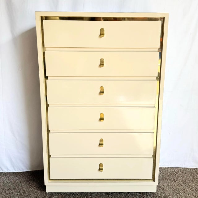 This striking postmodern highboy dresser features a glossy cream laminate finish framed by polished gold trim, exuding...