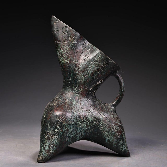 This is a cast bronze tripod vessel, featuring a distinctive form with a tall, pointed spout, a curved loop handle, and...