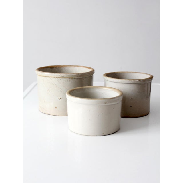 Set of three vintage stoneware crocks in graduated sizes, each with creamy white glaze, softly aged rims, and subtle...