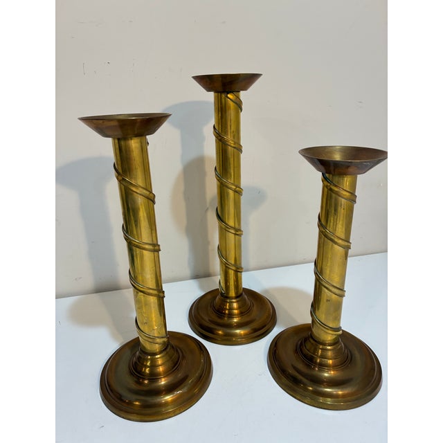 A striking vintage set of three graduated solid brass candlesticks featuring elegant spiral-wrapped columns and...
