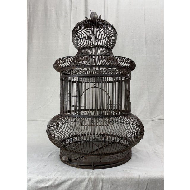 A striking architectural birdcage, in wrought iron with graceful scrollwork and tiered silhouette. The design evokes a...