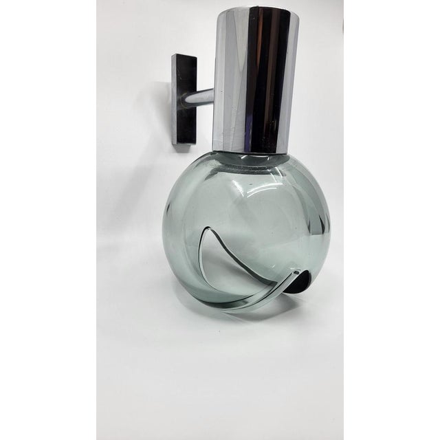 The eye-shaped wall light, made of smoked glass and chrome, perfectly embodies the Space Age style that marked the 60s....