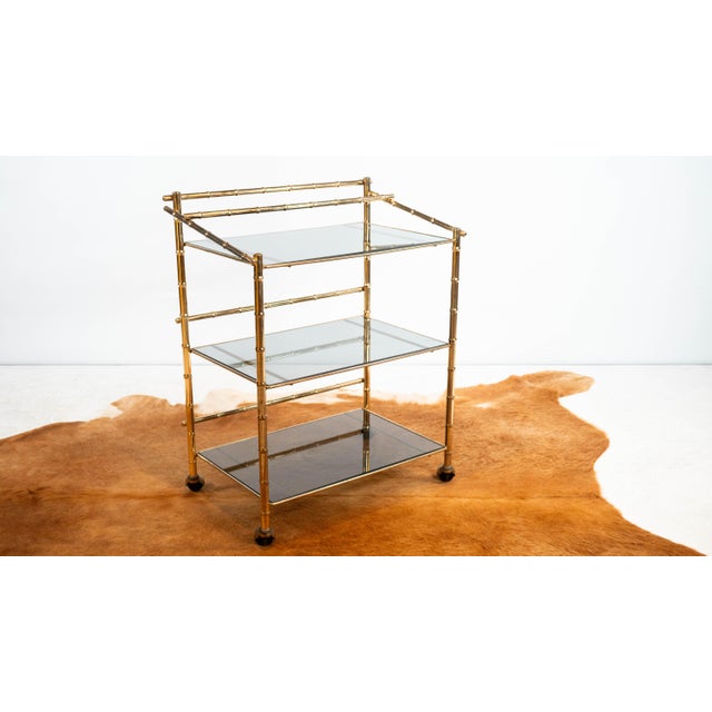 Vintage faux bamboo and smoked glass three-tier rolling bar cart, circa 1970s. Crafted from polished brass-finished metal...