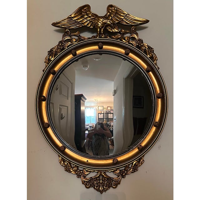 A Federal-style giltwood convex mirror crowned with a carved eagle, its wings outstretched above a laurel motif—an...