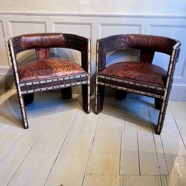 A striking pair of African hardwood and leather armchairs, richly decorated with carved detail, inlay and brass studwork....