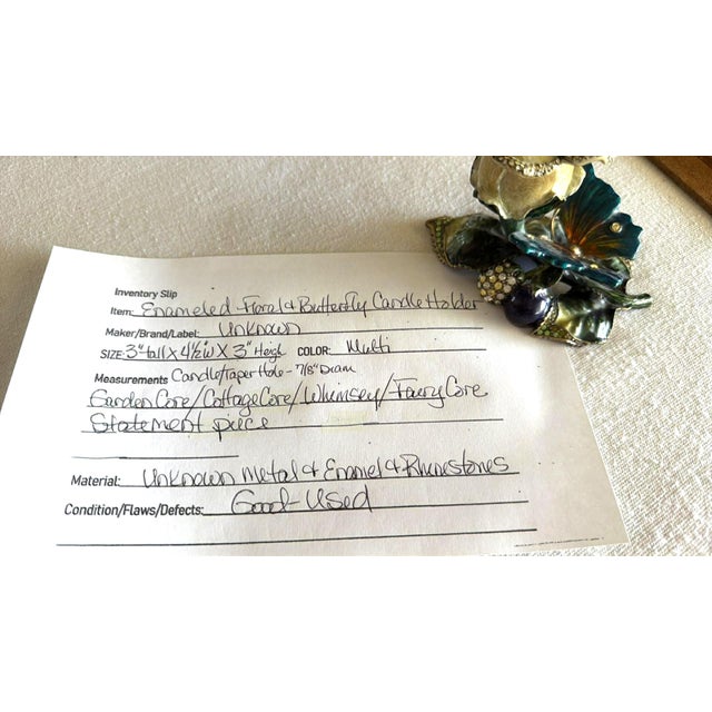 Ornate enameled metal candle holder features a botanical design with a cream-colored rose, vibrant teal butterfly, and...