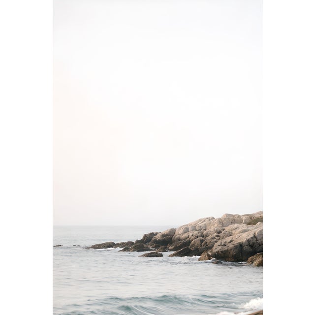 Minimalist coastal artwork featuring a white lighthouse set against soft ocean waves and rocky shoreline. The composition...
