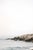Minimalist coastal artwork featuring a white lighthouse set against soft ocean waves and rocky shoreline. The composition...