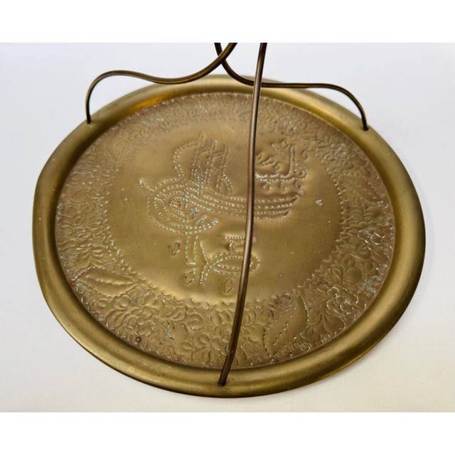 Round brass-tone serving tray with a raised floral border, ornate central decoration, and three curved supports meeting...
