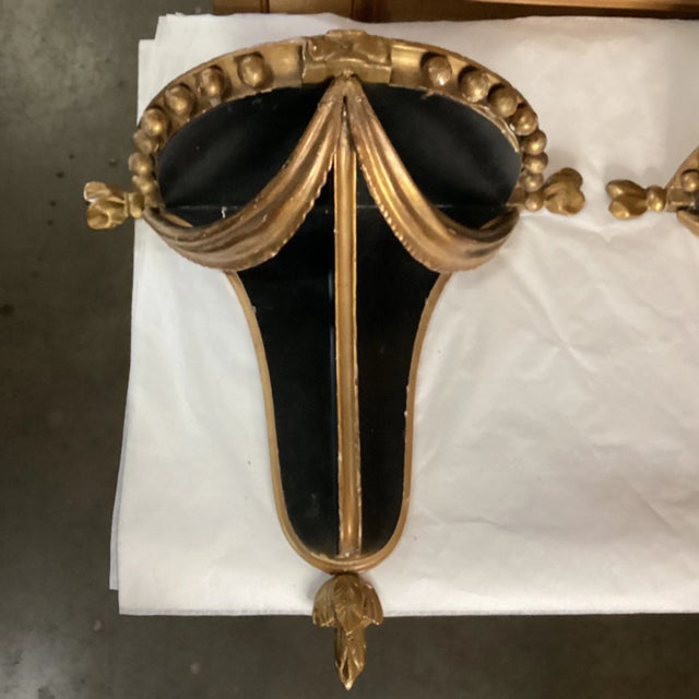 A pair of painted black and gold painted wood wall shelves. There are unique decorative elements that adorn the edges,...
