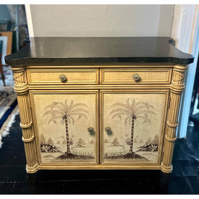 A sophisticated Hollywood Regency cabinet featuring a hand-painted tropical finish, carved bamboo-inspired corner pillars,...
