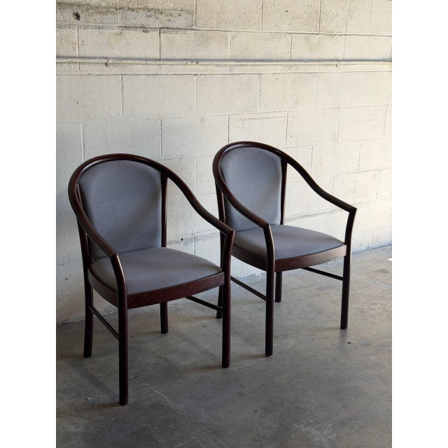 This pair of armchairs features a curved barrel frame with integrated armrests. Vintage 1980s. Crafted with dark mahogany-...