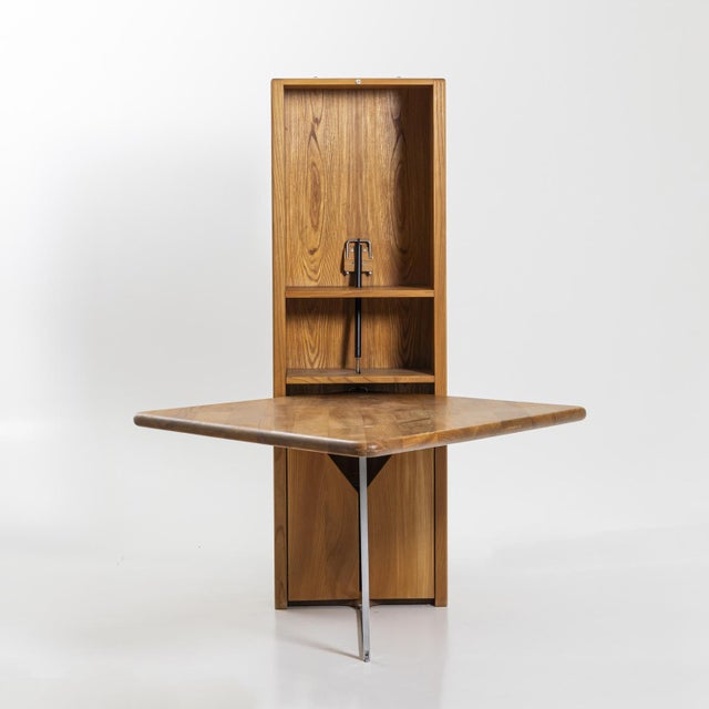 Narrow wooden cabinet with two doors and glass shelves in the lower section and a fold-down table top. The table top is...