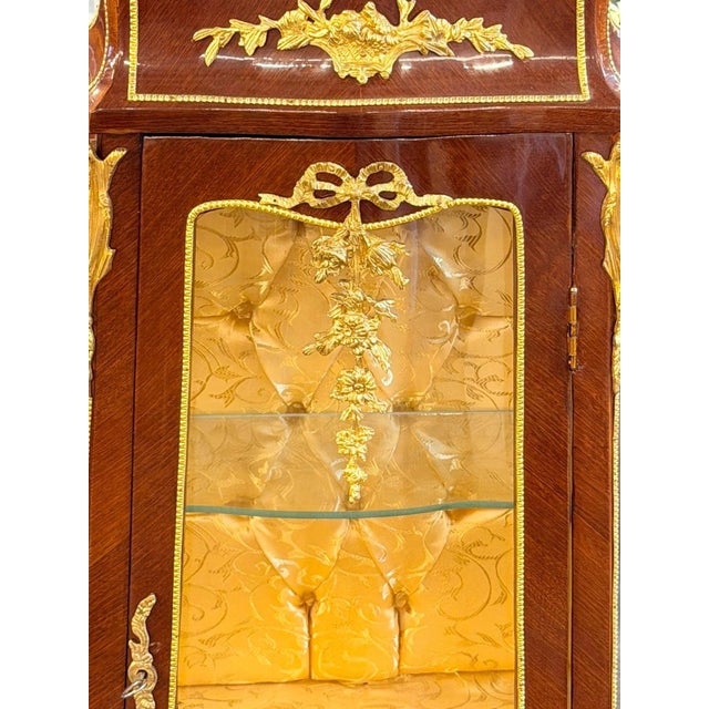 French Louis XVstyle petite vitrine cabinet, designed with a rich mahogany-toned finish, ornate gilt detailing, delicate...