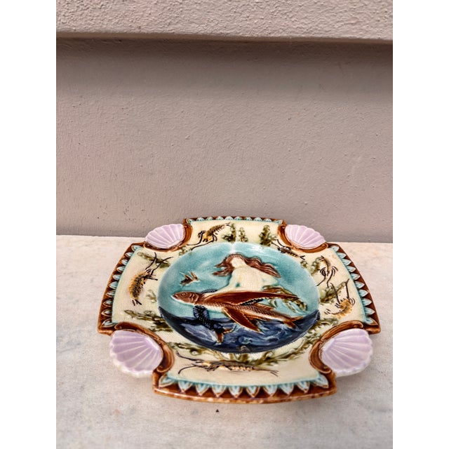Square Majolica mermaid plate with shells on the corners, the mermaid sit on a flying fish, the border is decorated with...