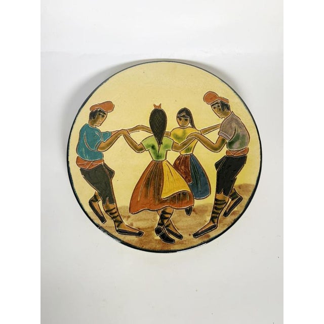 A charming round glazed ceramic dish featuring a figurative scene of four dancers in a circle, wearing stylized...
