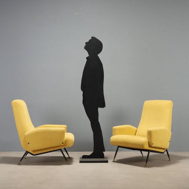 Two 1960s Italian-made armchairs with painted metal legs, brass tips, and a cushioned seat upholstered in yellow fabric....