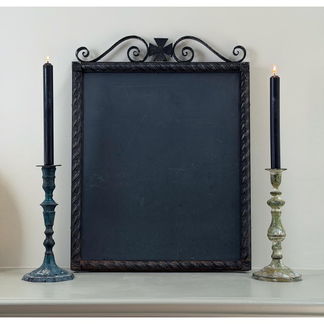 A salvaged slate chalkboard set in a dark iron frame, topped with a scrolled wrought iron crest and cross, and surrounded...