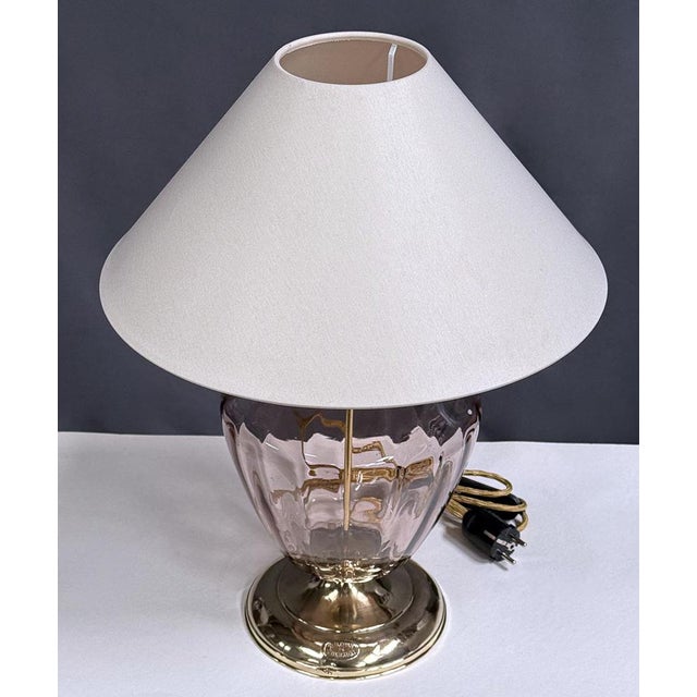 An elegant table lamp featuring a softly purple glass body and a shiny brass base. The vase-like shape of the glass...