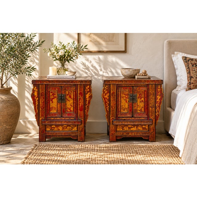 Two small cabinets in worn red lacquer stand side by side, their double doors painted with gilt decoration that has...