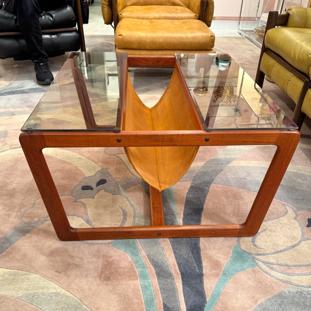 An elegantly sculpted teak coffee table with smoked glass top and cognac colored suede magazine sling. The frame is...
