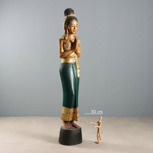 A polychrome and gold-painted wooden sculpture of a female figure in traditional Thai dress. The posture indicates the...