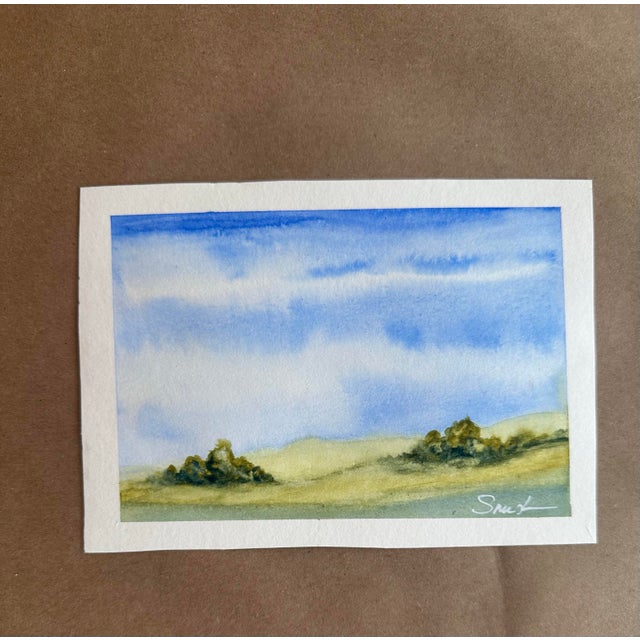 A peaceful miniature watercolor landscape featuring rolling hills, distant trees, and expansive blue skies rendered in...