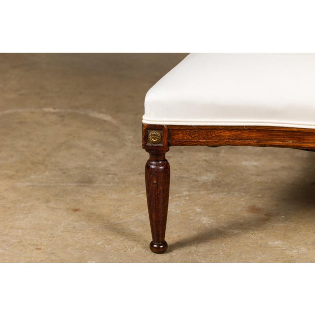 A French wooden ottoman from circa 1900 with tapering cylindrical legs, carved giltwood rosettes and muslin double welt...