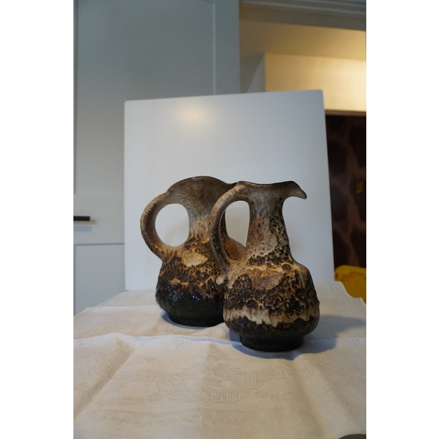 A small West German fat lava jug — cream-to-tan body under a thick dark volcanic glaze, with a pinched spout, a single...