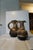 A small West German fat lava jug — cream-to-tan body under a thick dark volcanic glaze, with a pinched spout, a single...