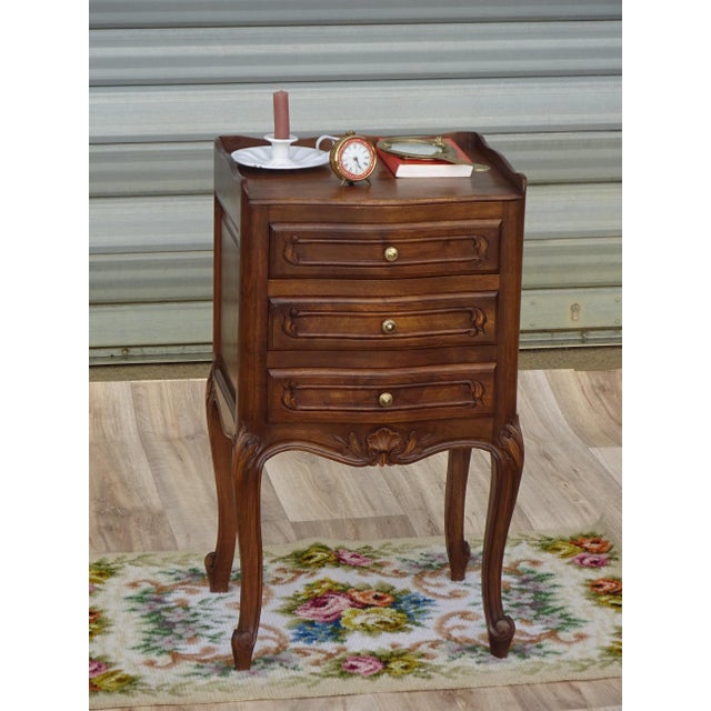 Elegant bedside table with three drawers and raised edges on three sides of the top. The tall, slender base features four...