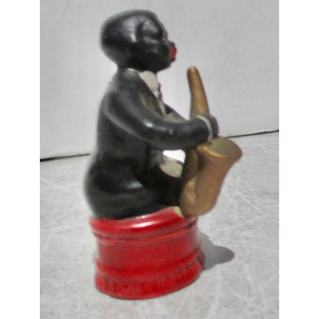 A ceramic figurine in the form of a black male musician playing the trumpet while sitting on a red ottoman. The musician...