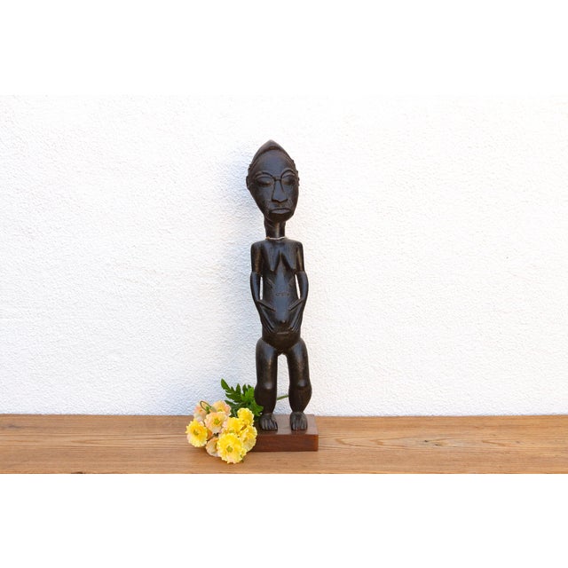 A slim standing woman carved in dark wood and polished to a deep, glossy black. She has an elongated, ringed neck, a close...