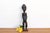 A slim standing woman carved in dark wood and polished to a deep, glossy black. She has an elongated, ringed neck, a close...