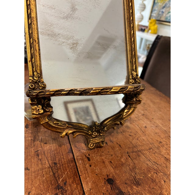 Antique giltwood small wall mirror with an integrated shelf, featuring an ornate design with a bow at the top. The slight...