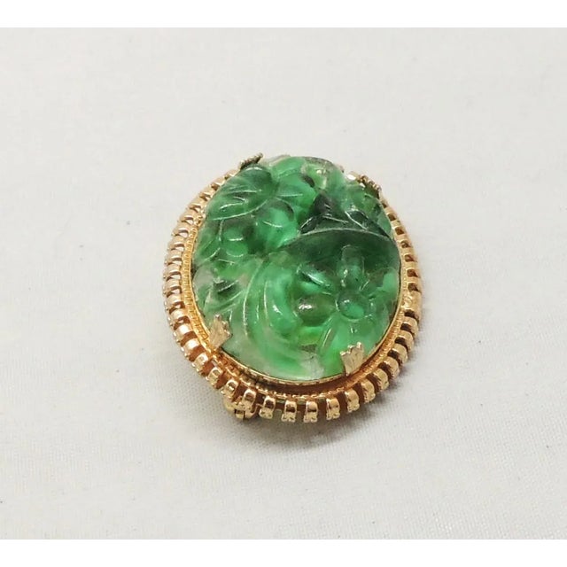 This is part of Chairish’s Costume Jewelry assortment. 1965 oval goldtone carved faux-jade brooch with security clasp....