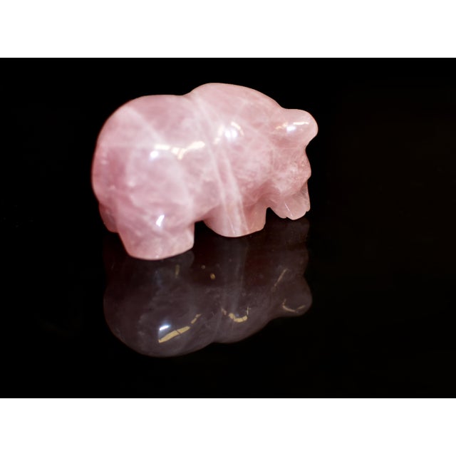 A charming hand-carved bear sculpture in natural rose quartz, radiating a soft, translucent blush tone with delicate...