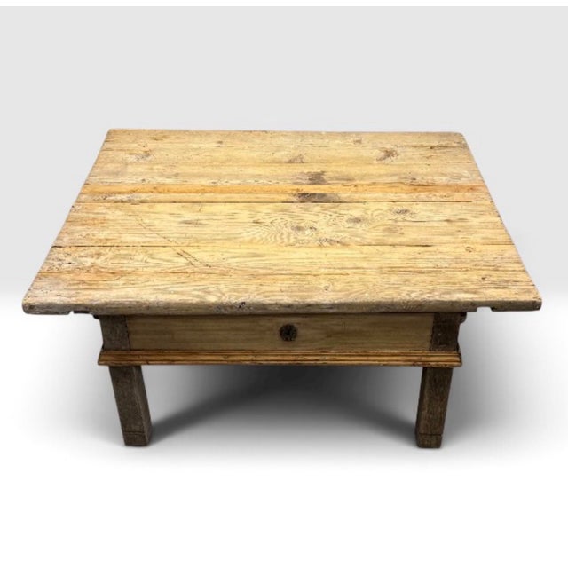 A rustic antique coffee table featuring a heavy plank top, single central drawer, and robust block legs. Originating from...